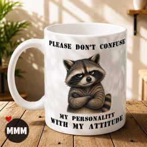 May include: A white ceramic mug featuring a graphic of a raccoon with crossed arms and a stern expression. The text printed on the mug reads, "PLEASE DON'T CONFUSE MY PERSONALITY WITH MY ATTITUDE," positioned above and below the raccoon illustration.