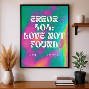Op de afbeelding: Een ingelijste kunstafdruk met de tekst "ERROR 404: LOVE NOT FOUND" in het wit, tegen een kleurrijke achtergrond van groen, roze en oranje tinten. Het kunstwerk wordt gepresenteerd op een houten oppervlak, met een vaas met gedroogde planten en een potplant in de buurt.