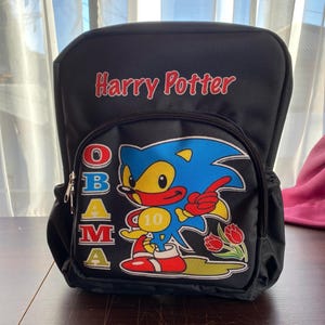 Puede incluir: Mochila negra con las palabras "Harry Potter" en letras rojas. El bolsillo delantero presenta un personaje de dibujos animados con pelaje azul, vientre amarillo y zapatos rojos. El nombre "OBAMA" está escrito verticalmente en el lado izquierdo.