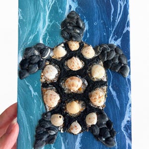 May include: A handcrafted sea turtle artwork made from seashells and dark pebbles. The turtle is set against a blue background with white wave-like patterns. The turtle's shell is composed of various seashells, creating a unique coastal decor piece.
