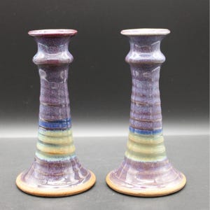 May include: A pair of ceramic candle holders featuring a glossy glaze with horizontal bands of purple, blue, and cream. Each holder has a flared base, a tapered cylindrical body, and a wide rim at the top for holding a candle securely.