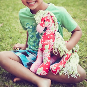 May include: A young person sits on green grass holding a handmade red and pink horse toy with green yarn hair. The toy has a floral pattern and a green button nose. The person is wearing a green t-shirt with a blue and black patterned shorts. The image includes the text "Gracie May Designs".