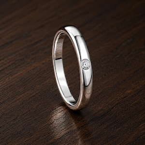 May include: A polished silver-toned band ring featuring a single, small, round, clear faceted gemstone set flush into the center of the outer surface. The ring has a smooth, rounded profile and a reflective metallic finish throughout its structure.