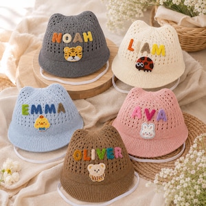 Puede incluir: Cinco sombreros de pescador tejidos con orejas de gato y correas para la barbilla. El sombrero gris tiene el nombre NOAH y un parche de tigre. El crema dice LIAM con una mariquita. El azul dice EMMA con un pollito. El rosa dice AVA con un conejo. El marrón dice OLIVER con un oso.