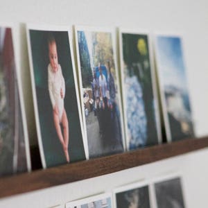 Peut inclure: Une collection de photographies encadr&eacute;es dispos&eacute;es sur une &eacute;tag&egrave;re. Les photos comprennent un b&eacute;b&eacute; en body blanc, un groupe de personnes marchant dans la rue, un gros plan d'une fleur et une sc&egrave;ne de paysage.