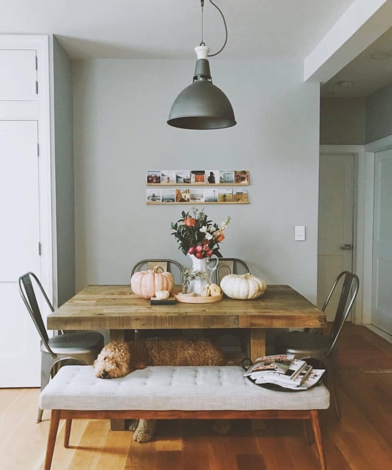 Peut inclure: Une table &agrave; manger en bois rustique avec un banc et des chaises. La table est d&eacute;cor&eacute;e d'un vase de fleurs, de citrouilles et d'un plateau d'objets d&eacute;coratifs. La pi&egrave;ce est peinte dans une couleur gris clair et a un style moderne de ferme.