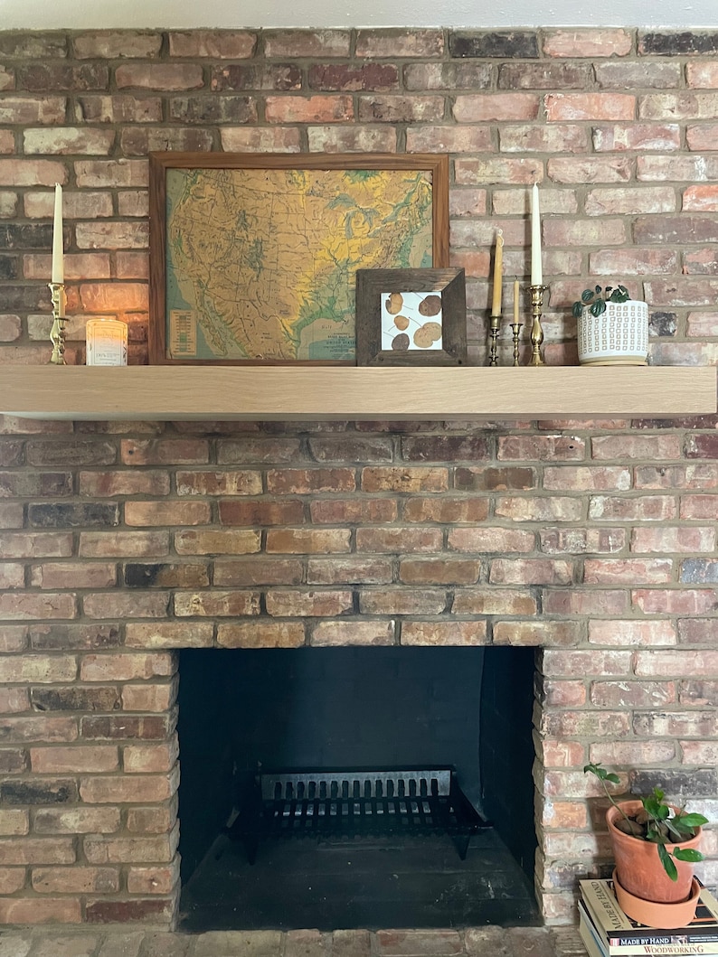 May include: A brick fireplace with a black metal grate and a wooden mantel. The mantel is decorated with a vintage map of the United States, a small framed print, candlesticks, and a potted plant.