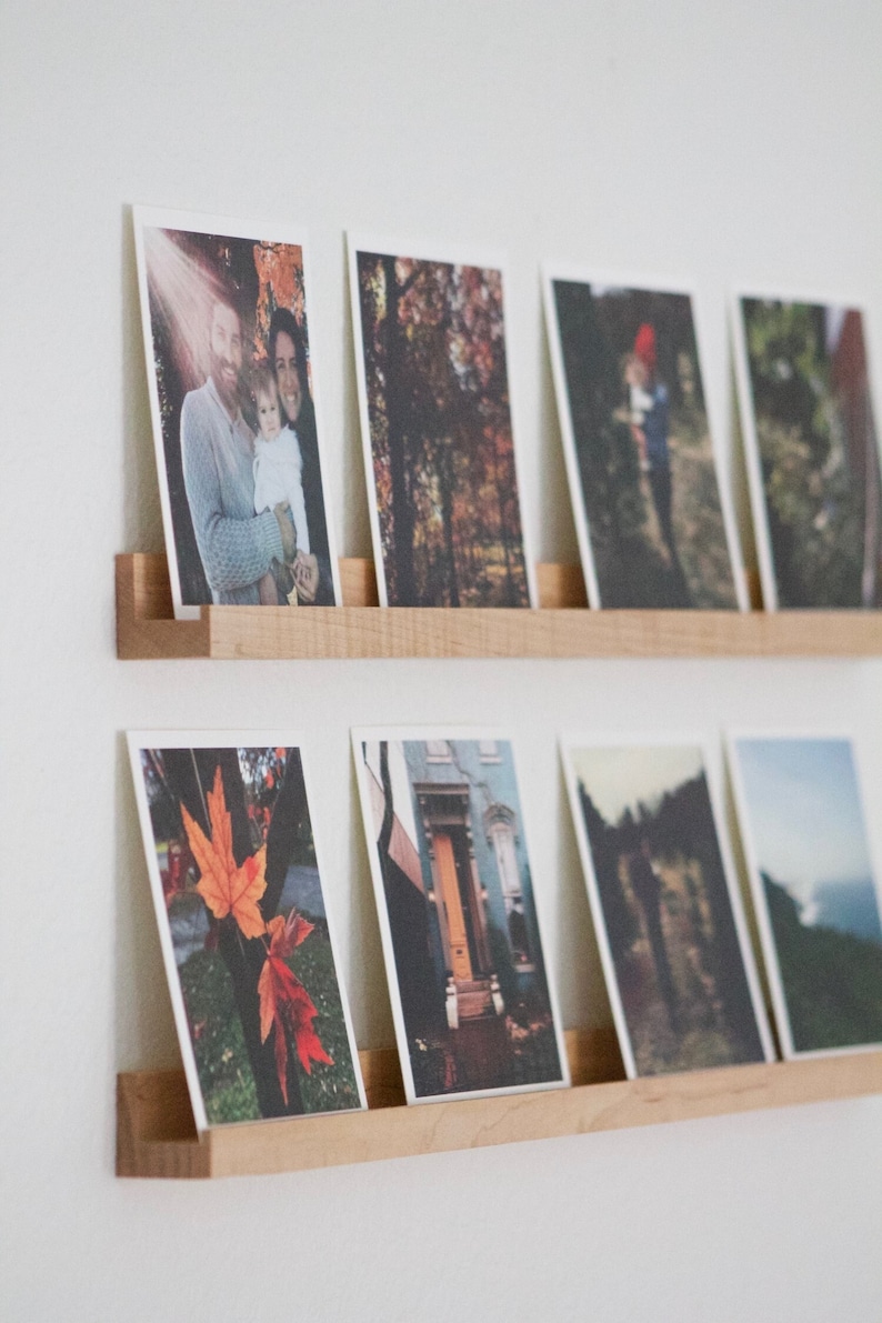 Peut inclure: Un ensemble d'&eacute;tag&egrave;res &agrave; photos en bois avec une vari&eacute;t&eacute; de photographies expos&eacute;es. Les &eacute;tag&egrave;res sont fix&eacute;es &agrave; un mur blanc. Les photos comprennent un portrait de famille, une vue d'une for&ecirc;t, un b&acirc;timent avec une porte et un paysage.
