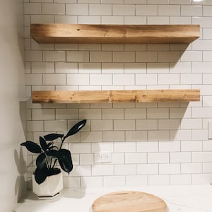 May include: Two wooden shelves mounted on a white tiled wall. A small potted plant sits on a white countertop below the shelves. A round wooden cutting board is on the countertop.