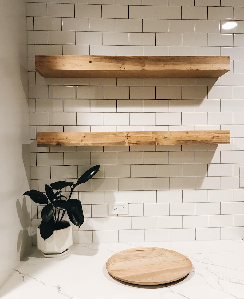 May include: Two wooden shelves mounted on a white tiled wall. A small potted plant sits on a white countertop below the shelves. A round wooden cutting board is on the countertop.