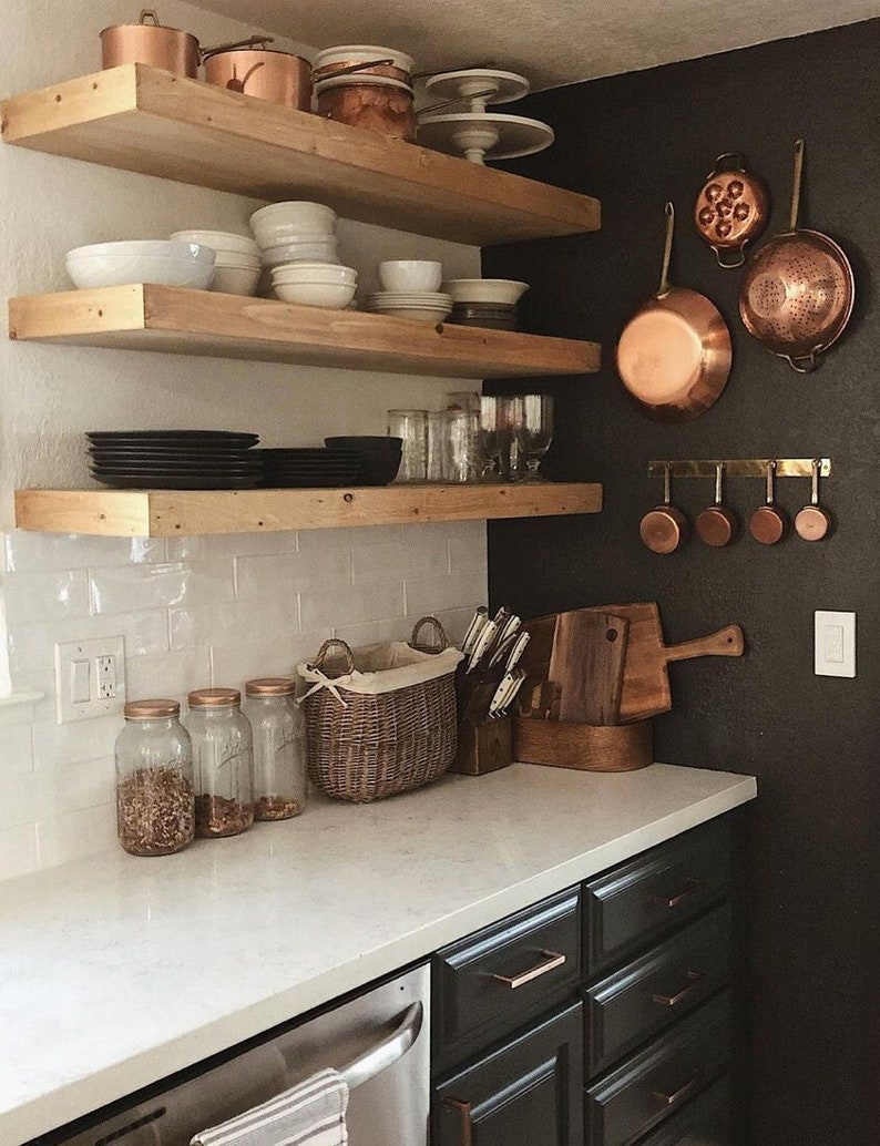 May include: A kitchen with white subway tile backsplash and dark cabinets. There are three wooden shelves with copper pots, bowls, and plates. On the wall, there are copper pots and measuring cups hanging on hooks.
