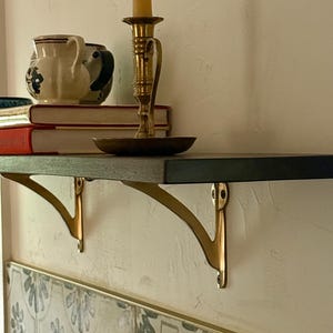 May include: A dark wooden shelf with brass brackets is mounted on a textured wall. A brass candlestick with a lit candle, books, and ceramic pitchers are displayed on the shelf. The shelf is above patterned tiles.