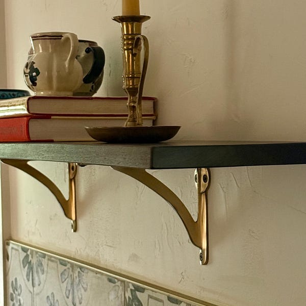 May include: A dark wooden shelf with brass brackets is mounted on a textured wall. A brass candlestick with a lit candle, books, and ceramic pitchers are displayed on the shelf. The shelf is above patterned tiles.