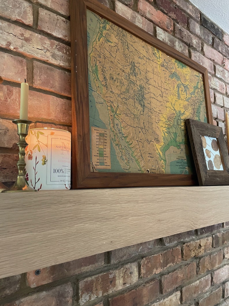 May include: A wooden mantel with a framed map of the United States, a candle holder with a lit candle, and a decorative candle jar with the text "Nature's Best" and "100% Soy Wax" on a brick wall background.