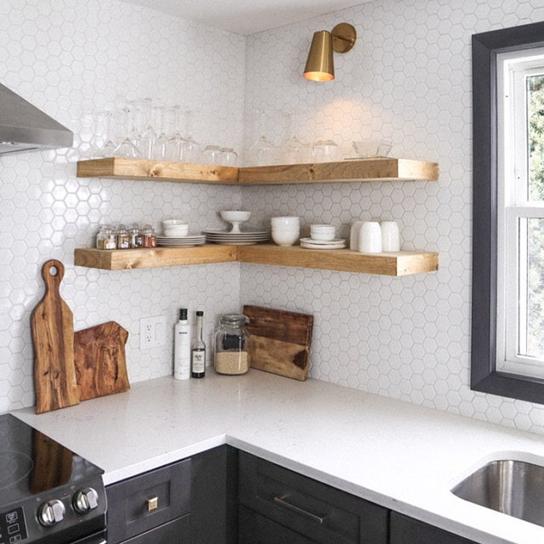 May include: Two wooden shelves mounted in a corner of a kitchen with white hexagonal tiles. The shelves are filled with dishes, glasses, and spices. A gold light fixture is mounted on the wall above the shelves.