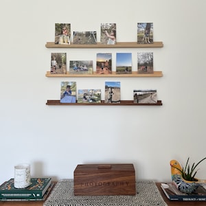Peut inclure: Trois &eacute;tag&egrave;res en bois avec une vari&eacute;t&eacute; de photographies encadr&eacute;es d'enfants en plein air. Sous les &eacute;tag&egrave;res se trouve une bo&icirc;te en bois avec le mot "PHOTOGRAPHIE" grav&eacute; sur le couvercle. La bo&icirc;te est pos&eacute;e sur une table avec un chemin de table &agrave; motifs noir et blanc.