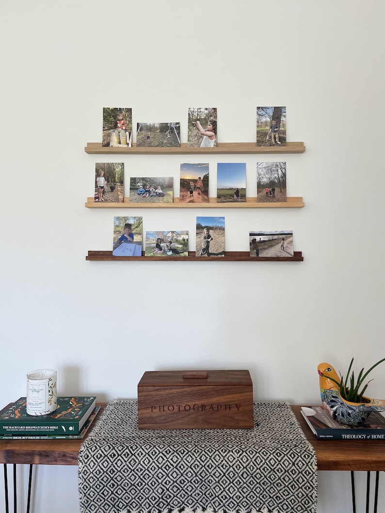 Peut inclure: Trois &eacute;tag&egrave;res en bois avec une vari&eacute;t&eacute; de photographies encadr&eacute;es d'enfants en plein air. Sous les &eacute;tag&egrave;res se trouve une bo&icirc;te en bois avec le mot "PHOTOGRAPHIE" grav&eacute; sur le couvercle. La bo&icirc;te est pos&eacute;e sur une table avec un chemin de table &agrave; motifs noir et blanc.