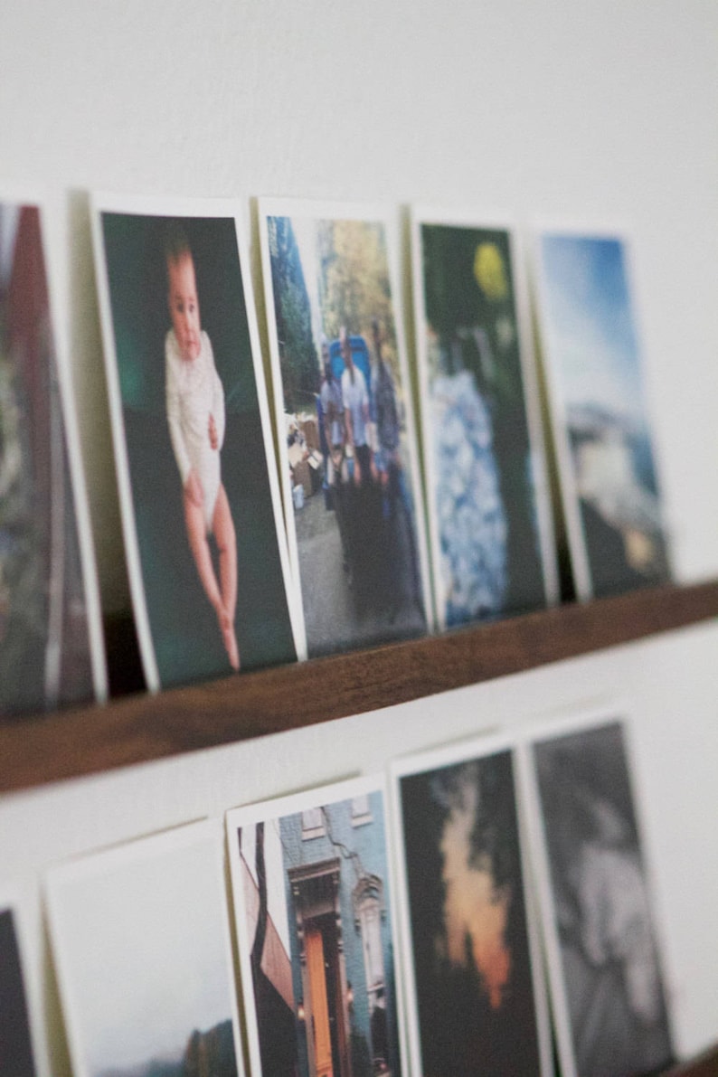 Peut inclure: Une collection de photographies encadr&eacute;es dispos&eacute;es sur une &eacute;tag&egrave;re. Les photos comprennent un b&eacute;b&eacute; en body blanc, un groupe de personnes marchant dans la rue, un gros plan d'une fleur et une sc&egrave;ne de paysage.
