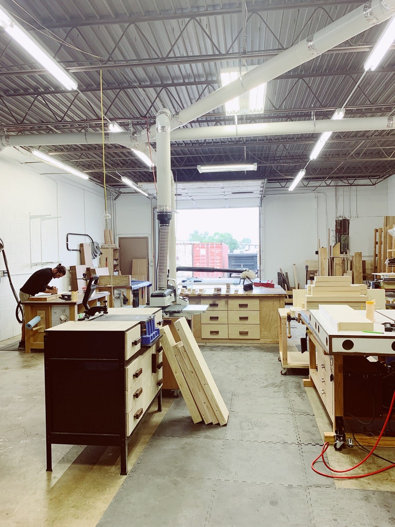 May include: A woodworking shop with a large workbench, tools, and wood pieces. The shop has a high ceiling with exposed beams and a large window.