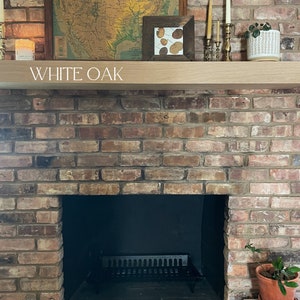 May include: A brick fireplace with a black metal grate and a wooden mantel. The mantel is labeled "WHITE OAK" and has a framed map of the United States, a framed picture with dried leaves, and candlesticks with candles.