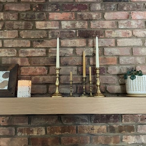 May include: A wooden shelf with a brick background. The shelf has a framed picture, a candle, and a potted plant. There are five gold candle holders with candles on the shelf.