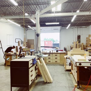 May include: A woodworking shop with a large workbench, tools, and wood pieces. The shop has a high ceiling with exposed beams and a large window.