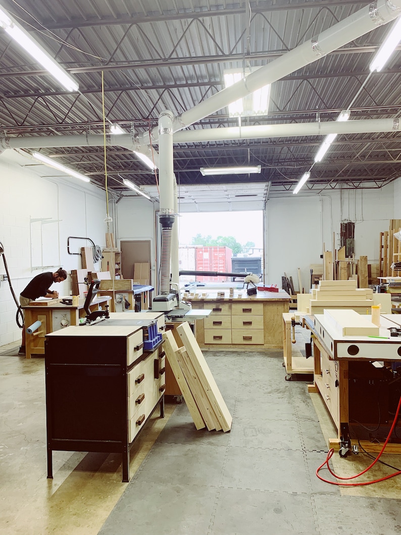 May include: A woodworking shop with a large workbench, tools, and wood pieces. The shop has a high ceiling with exposed beams and a large window.
