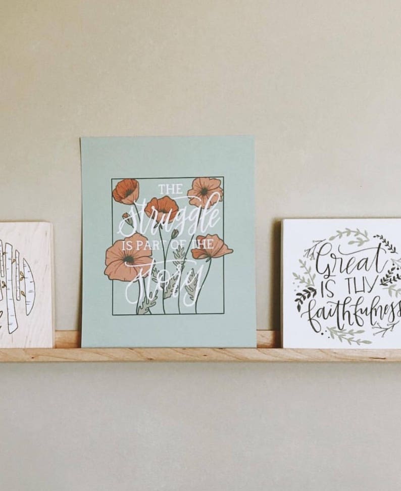 Peut inclure: Deux impressions encadr&eacute;es sur une &eacute;tag&egrave;re en bois. L'impression de gauche a un fond bleu clair avec une citation blanche qui dit "The struggle is part of the story" et pr&eacute;sente un motif floral avec des fleurs de pavot orange. L'impression de droite a un fond blanc avec une citation noire qui dit "Great is thy faithfulness" et pr&eacute;sente un motif floral avec des feuilles vertes.