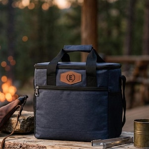 May include: A navy blue insulated cooler bag with black handles and a brown leather-like patch featuring the letter E in a circle. The bag has a zippered front pocket and a rectangular shape, sitting alongside a brown leather pouch and a metal mug.