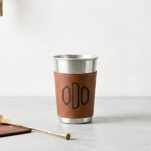 Könnte beinhalten: Ein Edelstahlbecher mit einer braunen Lederhülle, die ein dunkelbraunes, geprägtes Monogramm mit den Buchstaben ODO aufweist. Der Becher steht neben einem kleinen, dünnen, goldfarbenen Metalllöffel mit einem texturierten, gedrehten Griff.