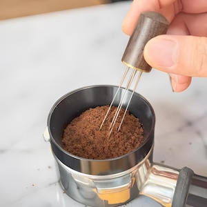 Könnte beinhalten: Ein Kaffee-Verteilerwerkzeug mit einem dunklen Holzgriff und dünnen Metallnadeln wird verwendet, um Kaffeepulver gleichmäßig in einem Siebträger zu verteilen. Der Siebträger ist schwarz und silberfarben und mit braunem Kaffeepulver gefüllt.