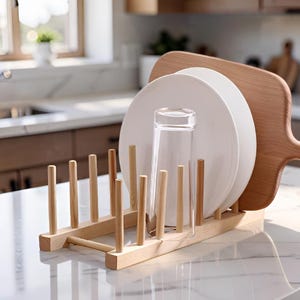 Peut inclure: Un égouttoir à vaisselle en bois clair doté de tiges verticales disposées en deux rangées parallèles. Le support contient deux assiettes en céramique blanche, un verre transparent et une planche à découper en bois clair avec poignée.
