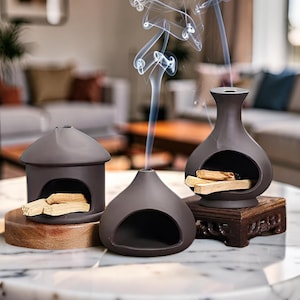 May include: Three dark brown ceramic incense burners with smooth, matte finishes. Two burners contain light-colored wooden sticks, while the center burner emits a thin, swirling trail of white smoke rising from its top opening.