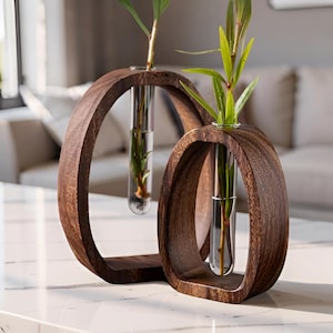 Peut inclure: Deux cadres en bois brun foncé de forme ovale, chacun contenant un vase en tube à essai en verre transparent. Chaque tube en verre contient de l'eau et une bouture de plante verte aux feuilles fines. Le bois présente un grain naturel.