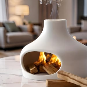 Puede incluir: Chimenea de mesa de cerámica blanca con una gran abertura ovalada que revela troncos de madera ardiendo y llamas. El humo se eleva desde la parte superior. La chimenea está sobre una superficie marmórea.