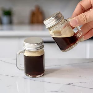 Puede incluir: Dos tazas de cristal en miniatura con tapas de rosca de color plateado, cada una llena de un líquido marrón oscuro cubierto con una espuma de color tostado claro. Las tazas de cristal transparente tienen un asa lateral y forma cilíndrica.