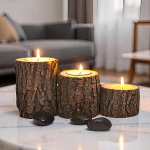 Può includere: Tre portacandele cilindrici realizzati con tronchi di legno naturale con corteccia marrone ruvida e strutturata. Ogni supporto contiene un lumino acceso con una calda fiamma gialla. Tre pietre di fiume lisce grigio scuro davanti.