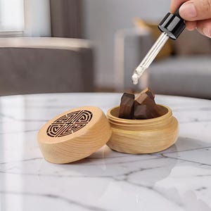 Peut inclure: Un récipient rond en bois clair avec un couvercle amovible présentant un motif géométrique découpé au laser. La base ouverte contient plusieurs petits blocs de bois brun foncé, et un compte-gouttes en verre est placé au-dessus.