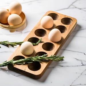 May include: A wooden egg holder with ten circular indentations, holding several brown eggs. A small woven basket with more eggs sits to the left. Sprigs of rosemary are placed around the holder. The holder is on a white marble surface.