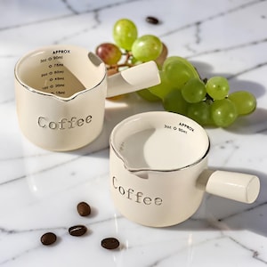 Op de afbeelding: Twee crèmekleurige keramische koffie-maatbekers met handvatten. De bekers hebben een zilveren rand en het woord "Coffee" is in reliëf op de voorkant aangebracht. Metingen zijn aan de binnenkant afgedrukt. Druiven en koffiebonen staan ook op de foto.