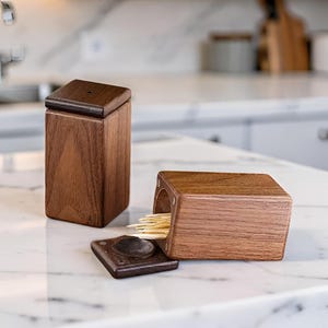 Peut inclure: Deux porte-cure-dents rectangulaires en bois de noyer brun foncé. L'un est posé verticalement avec son couvercle fermé, tandis que l'autre est couché sur le côté, couvercle ouvert, laissant apparaître un petit faisceau de cure-dents en bois clair.