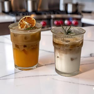 Puede incluir: Dos vasos de cristal transparente con bebidas heladas. El vaso izquierdo contiene una bebida de capas naranja y marrón con una rodaja de cítrico seco. El vaso derecho tiene una textura estriada y contiene café helado con capas blancas y marrones.
