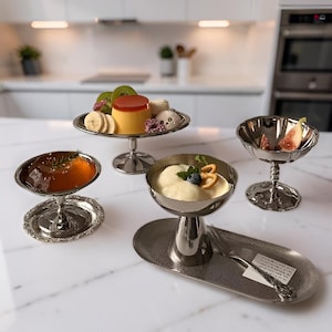 Peut inclure: Un ensemble de quatre plats de service en métal argenté, comprenant trois coupes sur pied et un bol peu profond, garnis de divers desserts comme de la crème, des fruits et de la gelée. Une petite fourchette repose sur un plateau ovale.