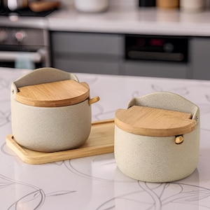 Peut inclure: Deux récipients ronds en céramique gris clair moucheté avec des couvercles en bois à charnières et de petites cuillères en bois. Un récipient repose sur un plateau rectangulaire en bois, tandis que l'autre est posé à côté.