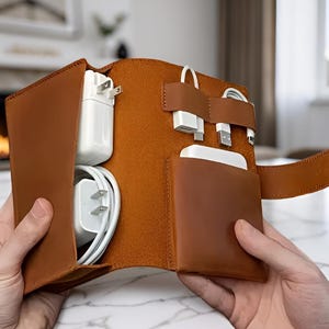 Peut inclure: Un étui organisateur en cuir havane doté de multiples fentes et poches intérieures conçues pour ranger des adaptateurs secteur blancs, des câbles de charge et une batterie externe rectangulaire, avec un rabat pour sécuriser le contenu.