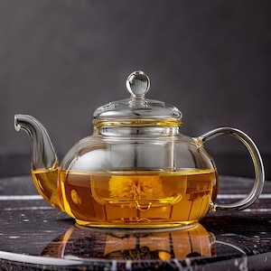 May include: A clear glass teapot with a curved spout and handle. The teapot is filled with amber-colored liquid and a yellow flower. The lid has a small, round knob. The teapot sits on a dark, patterned surface.