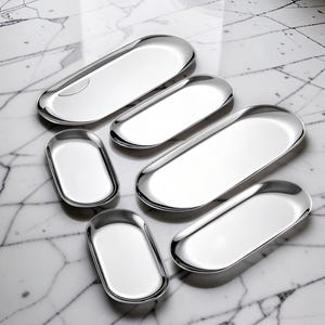 May include: A collection of six polished silver-toned metal trays in varying sizes, each featuring an elongated oval shape with smooth, rounded edges and a reflective, mirror-like surface finish, arranged in a cluster on a white surface.