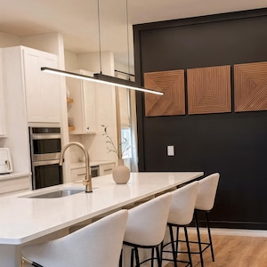 Op de afbeelding: Moderne keuken met een wit aanrecht eiland en barkrukken. Een zwarte lineaire hanglamp hangt boven het eiland. De keuken heeft witte kasten, een gouden kraan en decoratieve houten wandpanelen.