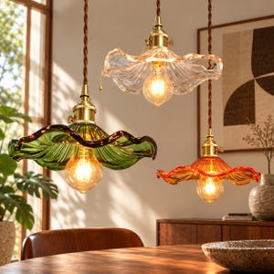 May include: Three pendant lights with ruffled, flower-shaped glass shades in clear, green, and amber colours. Each lamp features a brass-toned metal socket, a warm-toned Edison-style light bulb, and a brown twisted fabric cord for suspension.