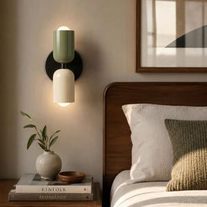 May include: A wall-mounted light fixture featuring two cylindrical shades in sage green and cream, attached to a circular black base. The fixture emits a soft glow from the top and bottom, illuminating the wall with a warm, focused light.